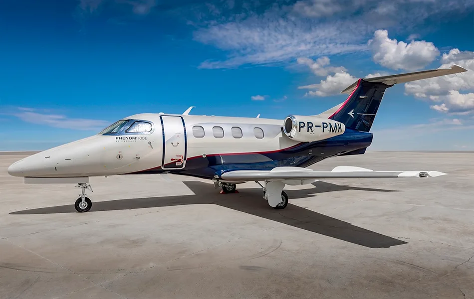 A white and blue Embraer Phenom 100E business jet with registration PR-PMX is parked on a tarmac under a partly cloudy sky.