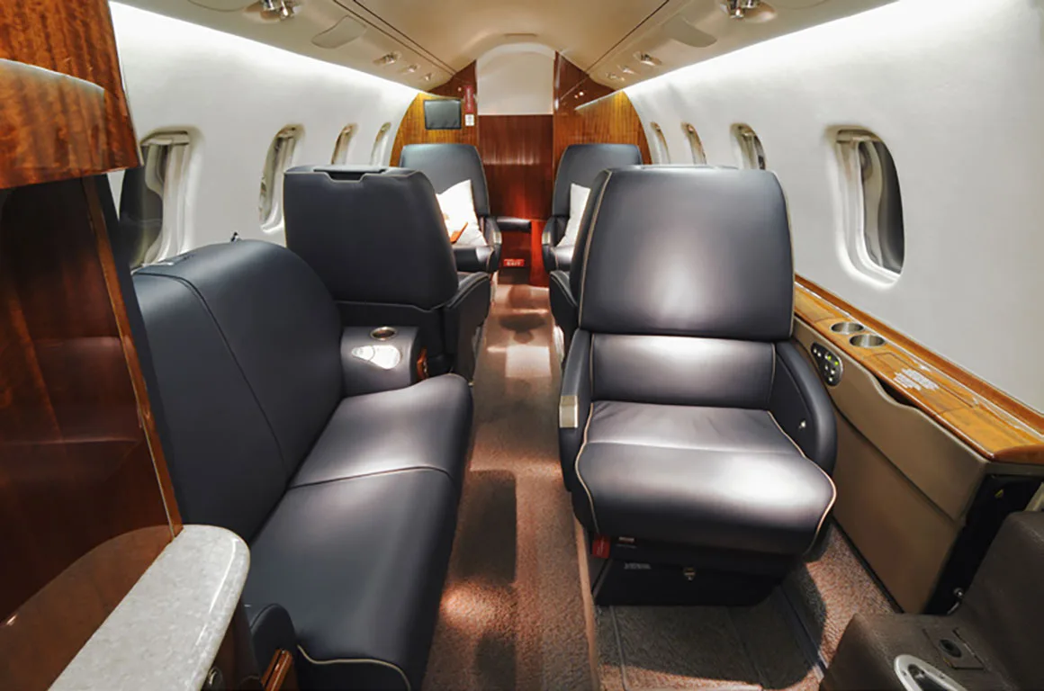 A view down the aisle of a private jet cabin, showing multiple black leather seats and rich wood paneling.