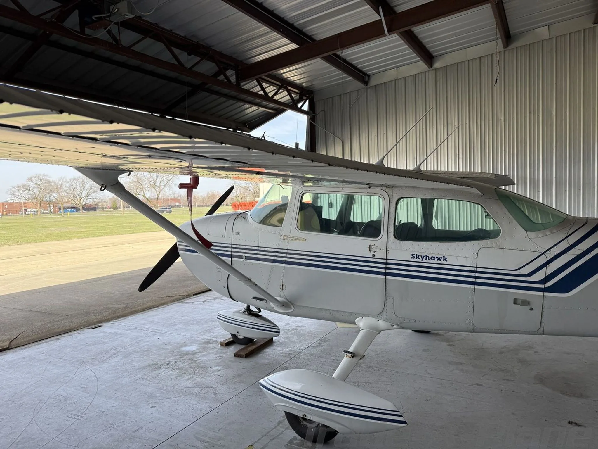 A white Cessna Skyhawk with blue stripes parked inside a hangar, viewed from the front-left, with an airfield visible outside.