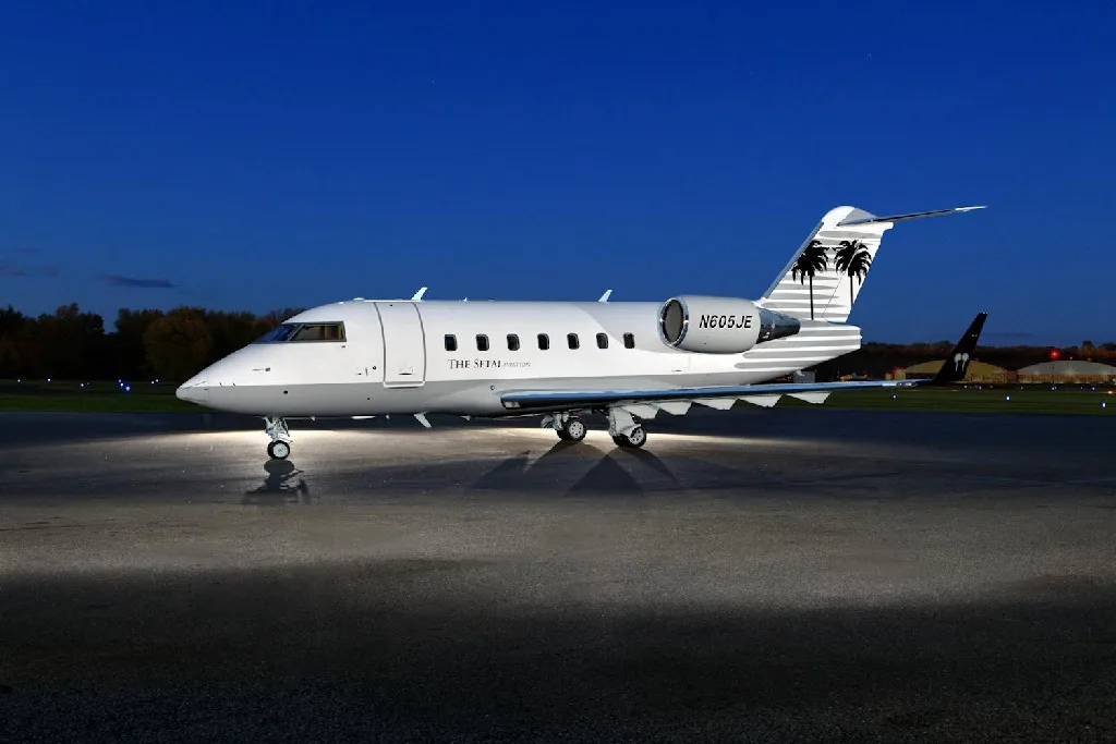 A white Bombardier Challenger 605 business jet, registration N605JE, with palm tree tail art, sits on a tarmac at night.