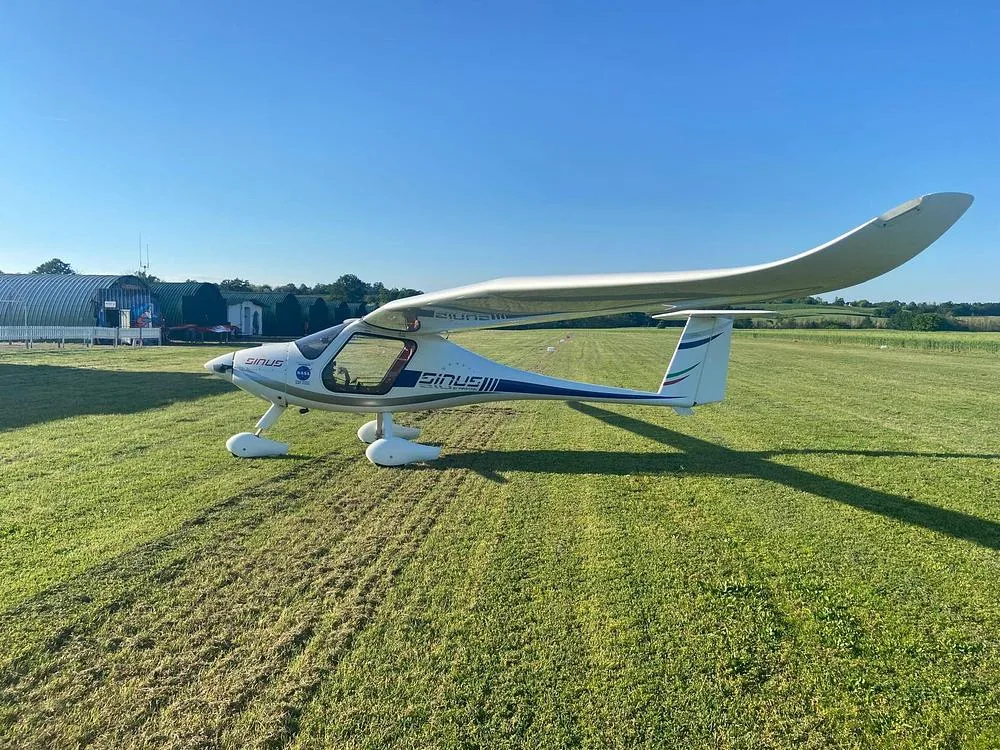A white Pipistrel Sinus aircraft with blue and green tail stripes, viewed from the side on a bright green airfield.