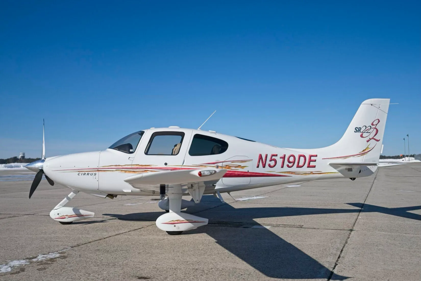 A white Cirrus SR22 aircraft with red and gold decals, registration N519DE, parked on a tarmac under a clear blue sky.