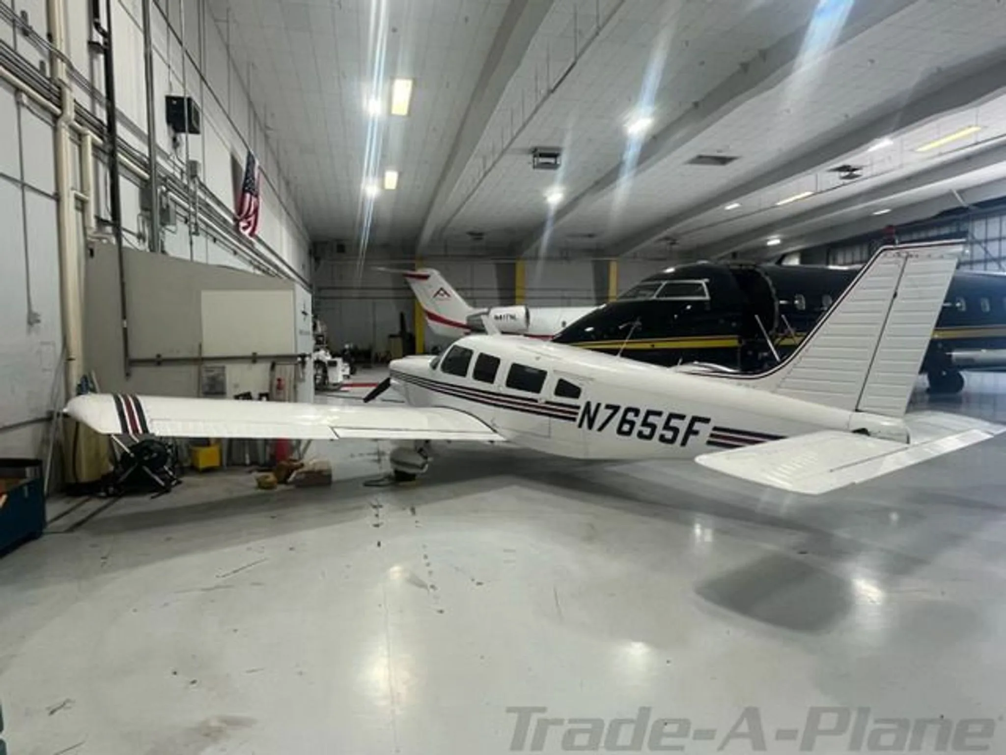 A white single-engine propeller aircraft with N7655F registration and red and black stripes is parked in a hangar, with other aircraft in the background.