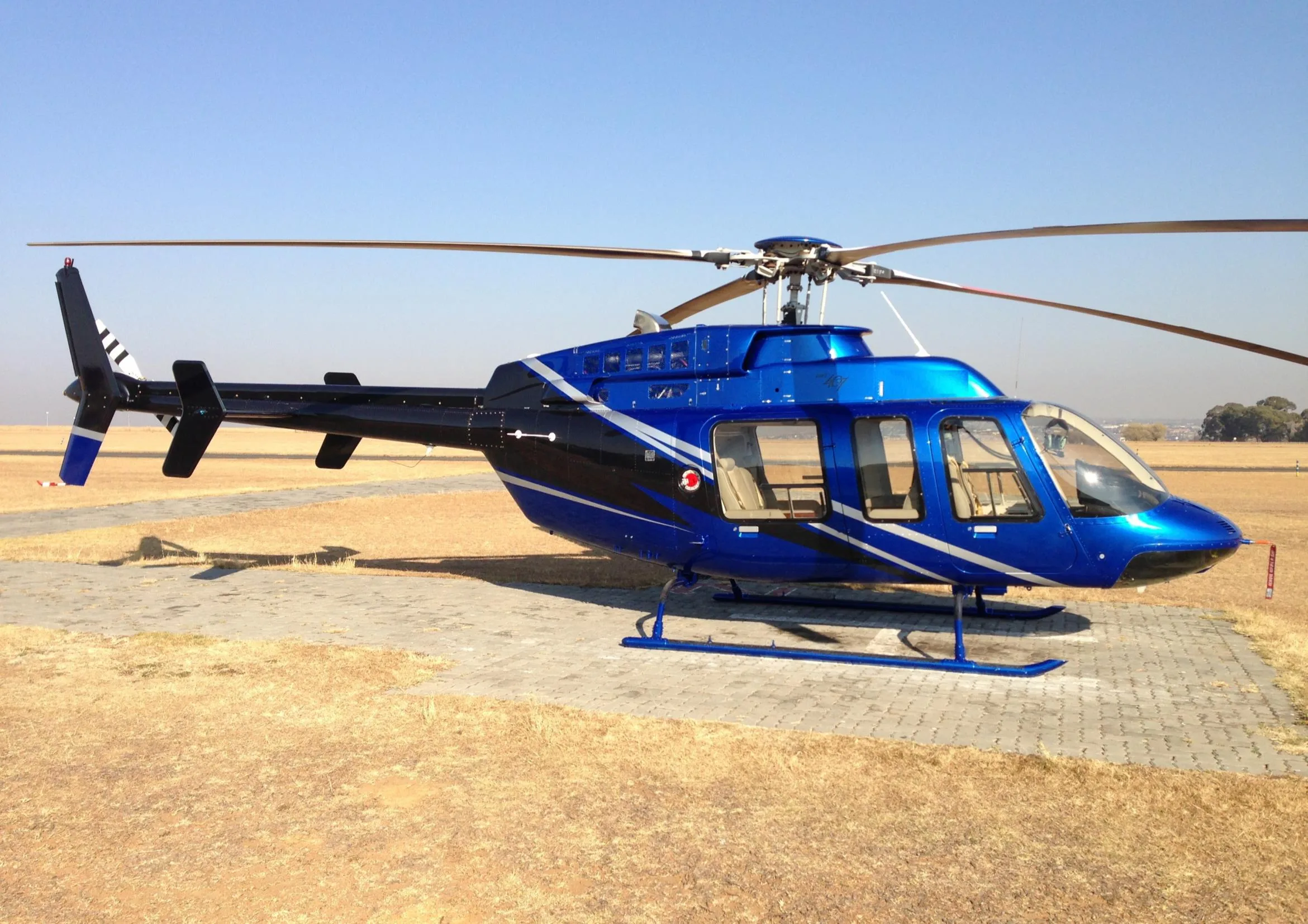 A blue and black helicopter with white stripes is parked on a paved area in a dry, grassy field under a clear sky.