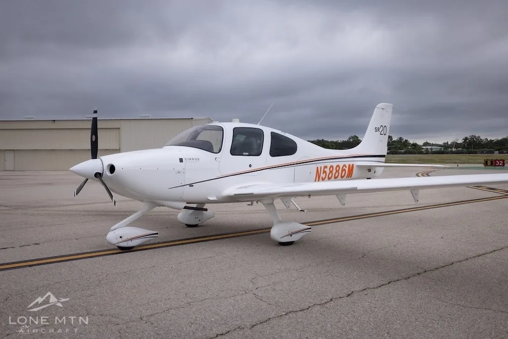 A white Cirrus SR20 aircraft with orange and black stripes, registration N5886M, parked on an airfield under a cloudy sky.