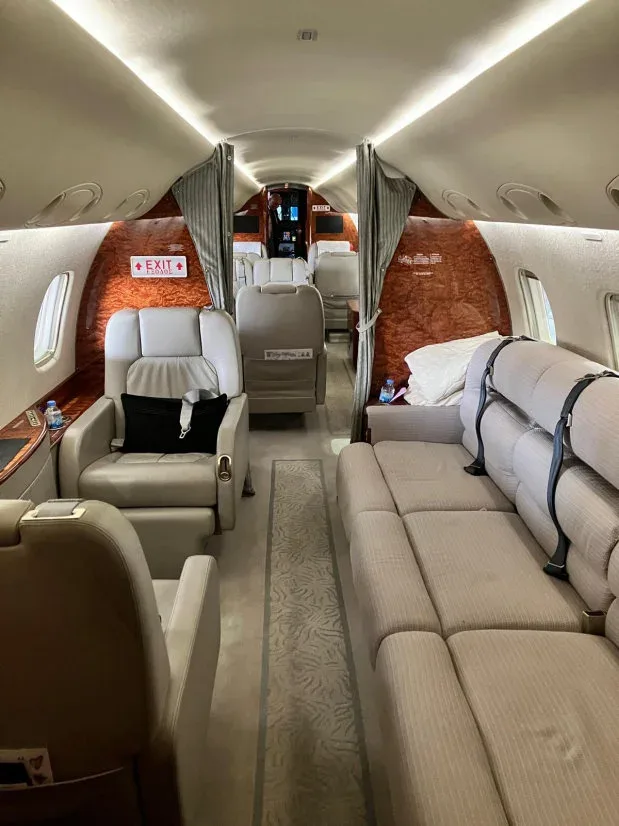 The luxurious interior cabin of a private jet features light grey leather seats, a long couch, and wood paneling.