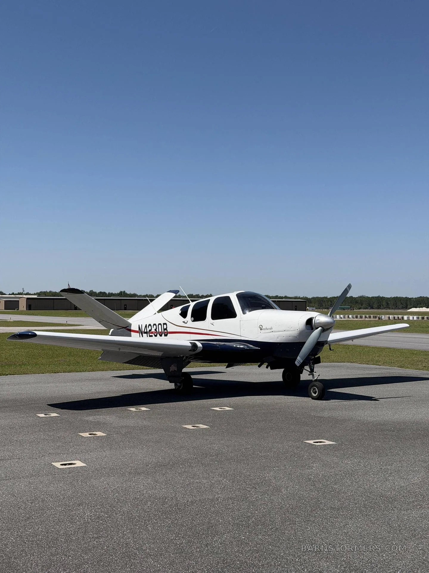 A white and blue Beechcraft Bonanza (N4230B) is parked on an airfield tarmac under a clear blue sky.