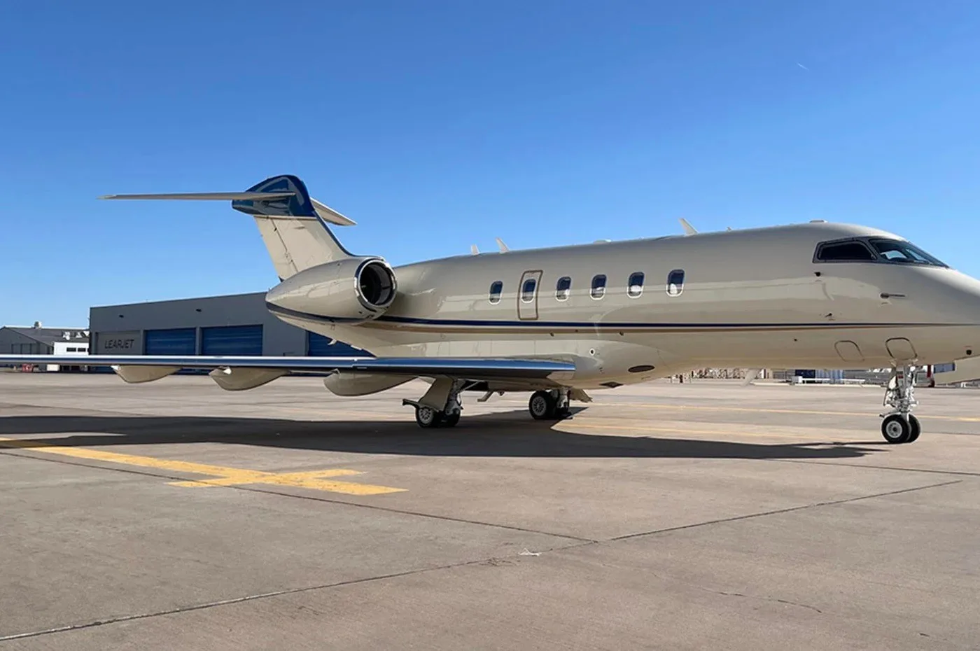 A beige and blue business jet, possibly a Learjet, parked on an airport tarmac next to a hangar.