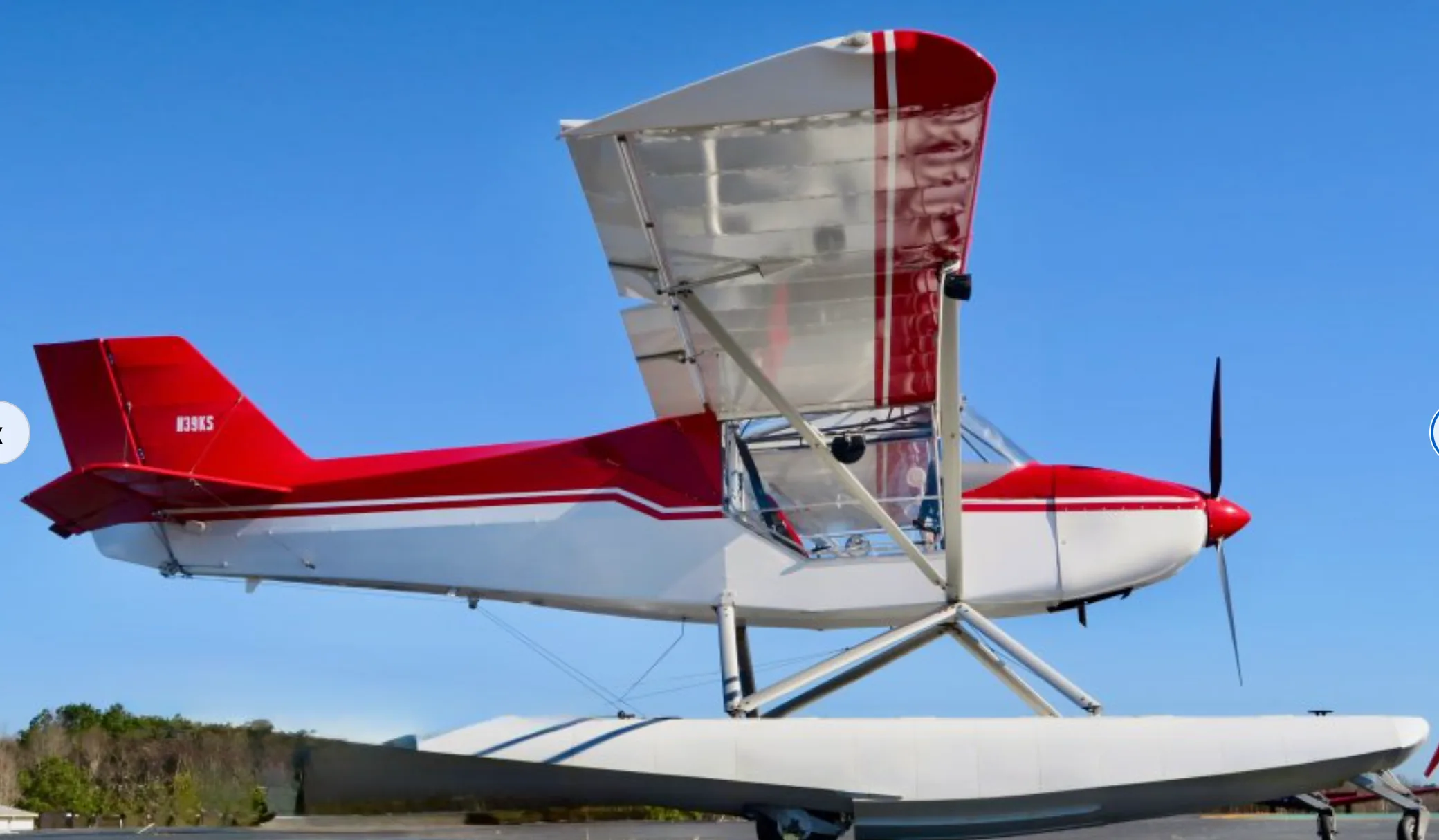 A side view of the red and white N39KS floatplane with its wings slightly angled upwards, parked on an airfield.