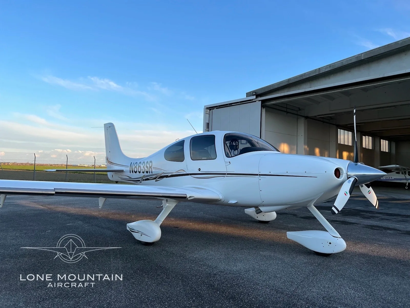 A white Cirrus SR22 aircraft with registration N863SR is parked in front of an open hangar at sunset.