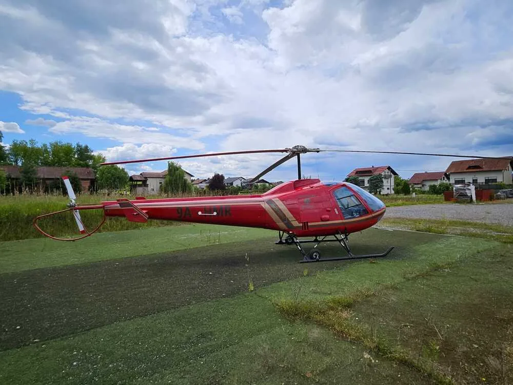 A red helicopter with gold stripes is parked on a green helipad, with houses and trees in the background under a cloudy sky.