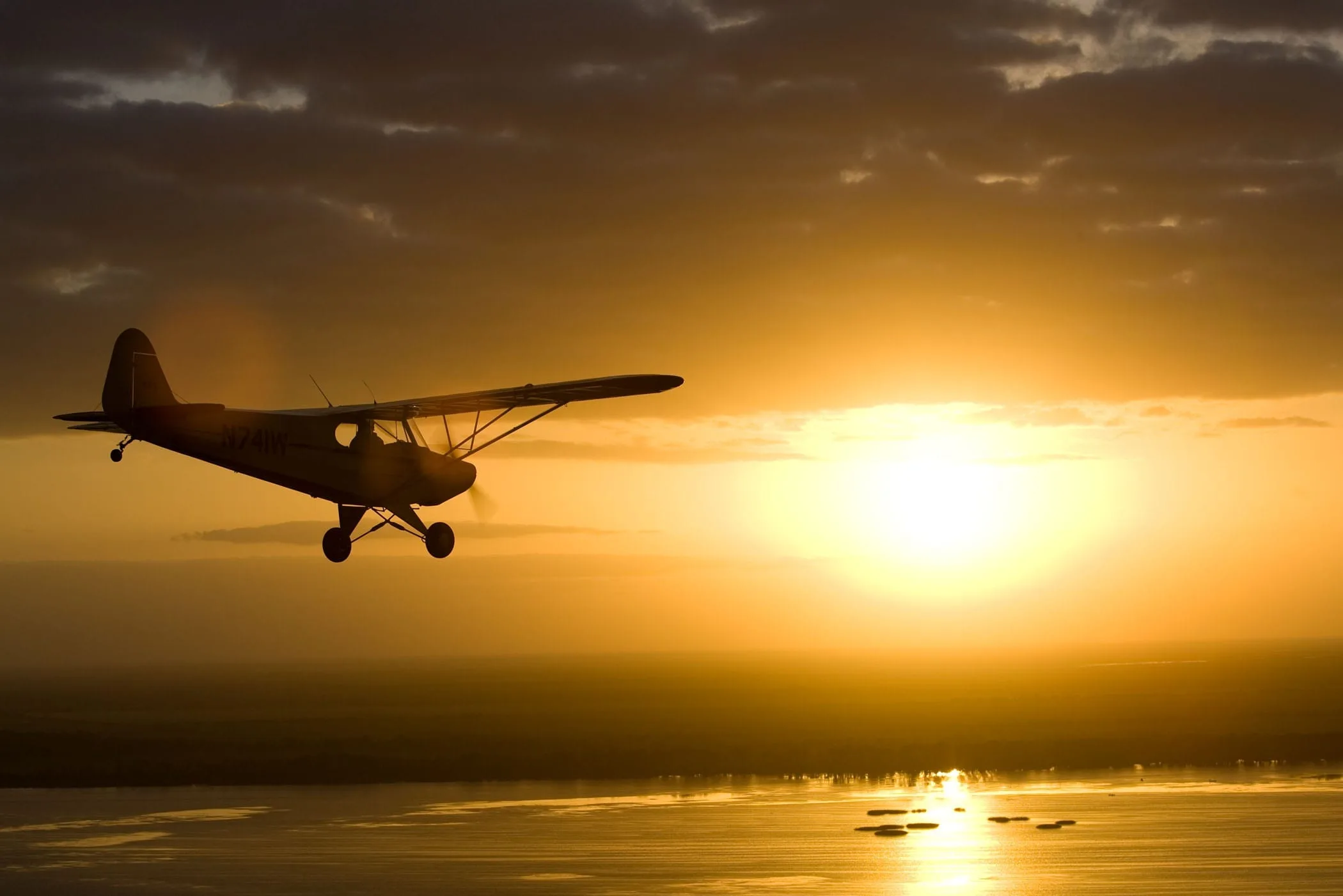 A yellow Piper Cub-style aircraft (N741W) is silhouetted against a bright orange sunset over a body of water.