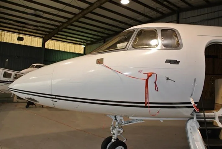 The white nose and cockpit area of a business jet with red 'REMOVE BEFORE FLIGHT' tags, parked inside a hangar.