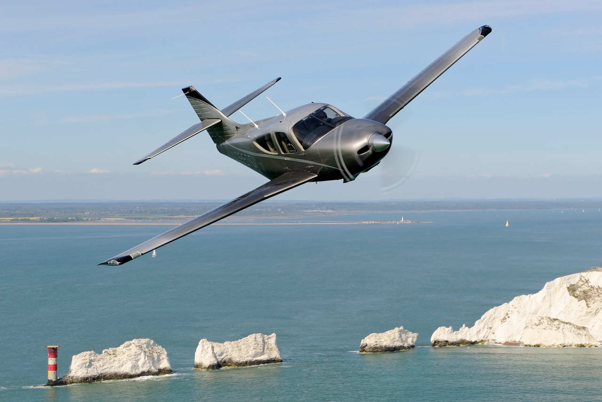 A dark grey single-engine propeller aircraft banks over the sea, with white cliffs and a lighthouse in the background.