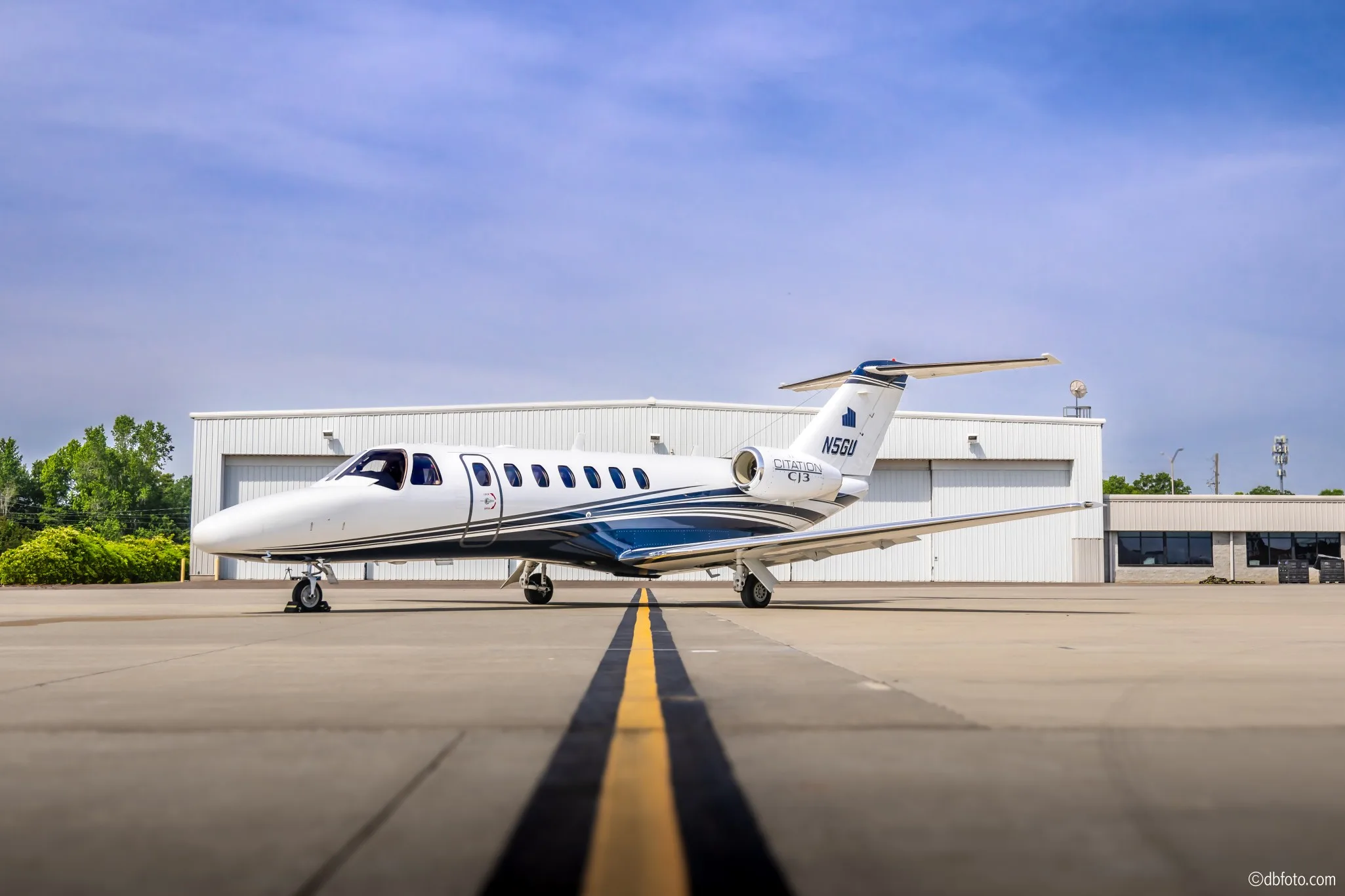A white and blue Cessna Citation CJ3 (N5GU) business jet is parked on an airport tarmac in front of hangars.