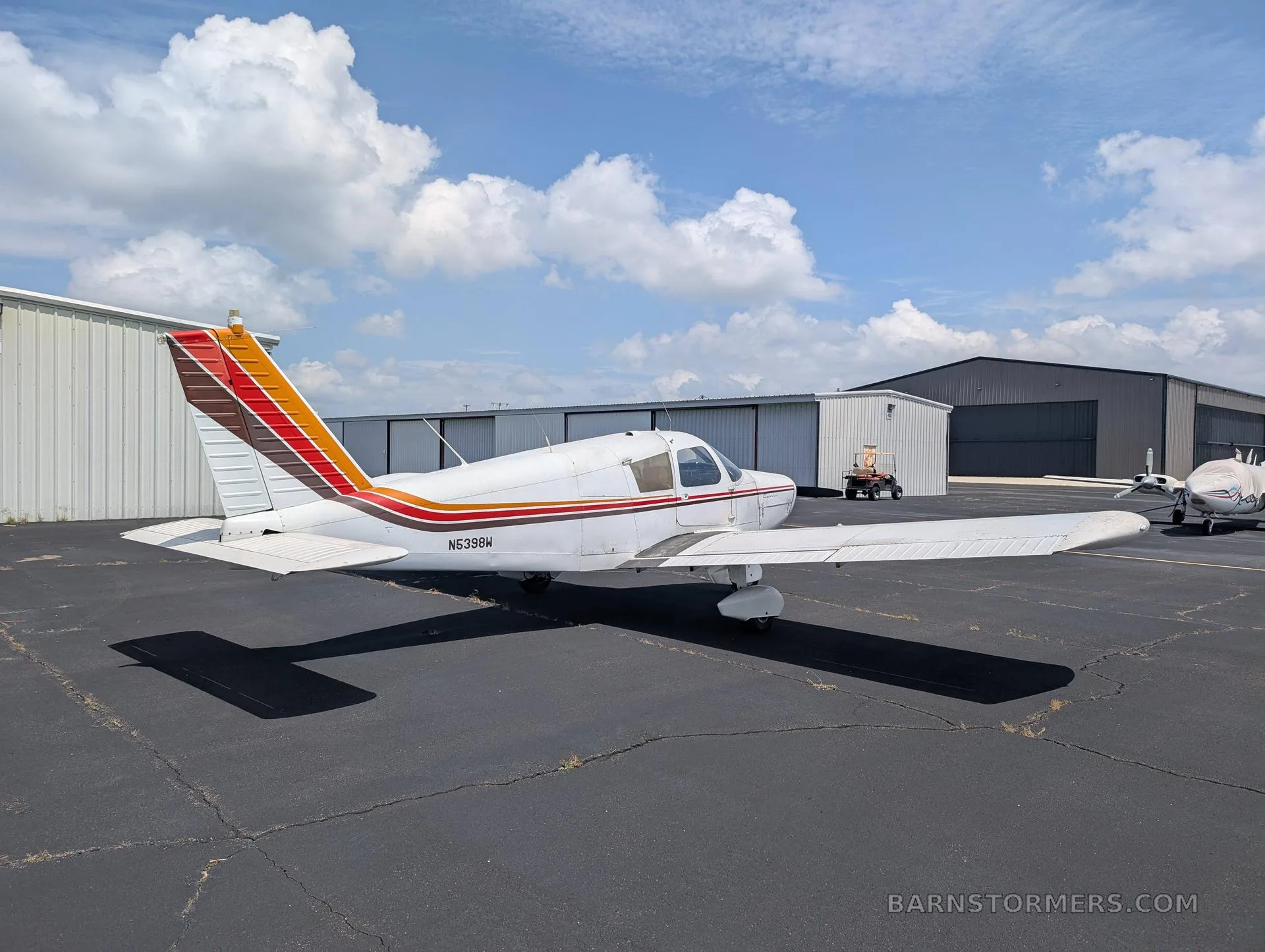 A white single-engine low-wing aircraft with red, orange, and brown stripes on the tail and fuselage, viewed from the left side, parked on an asphalt ramp.
