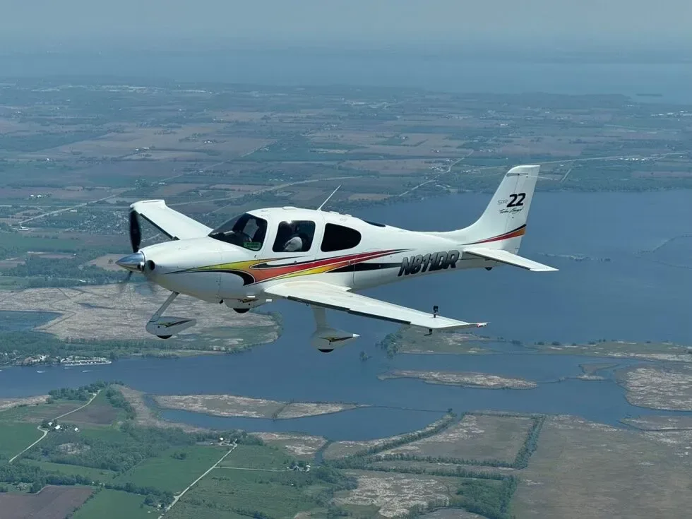 An aerial view of a white Cirrus SR22 aircraft with orange and red stripes flying over a lake and land.