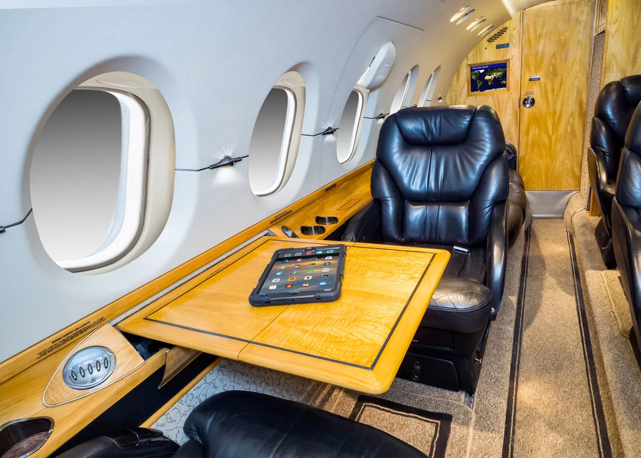 A private jet interior showing a black leather seat, a wooden table with a tablet, and large windows.