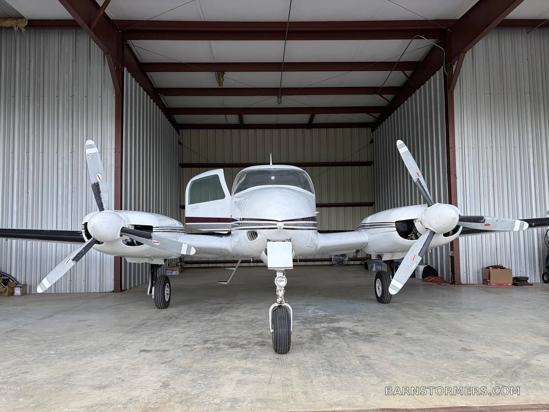 A white twin-engine propeller aircraft with dark stripes is parked inside a hangar, viewed from the front.