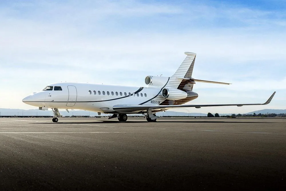 A Dassault Falcon 900EX business jet with white and brown livery is parked on an airfield under a blue sky.