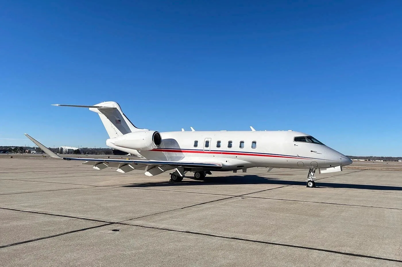 A white business jet with red and blue stripes on the fuselage, parked on a concrete tarmac under a clear blue sky.