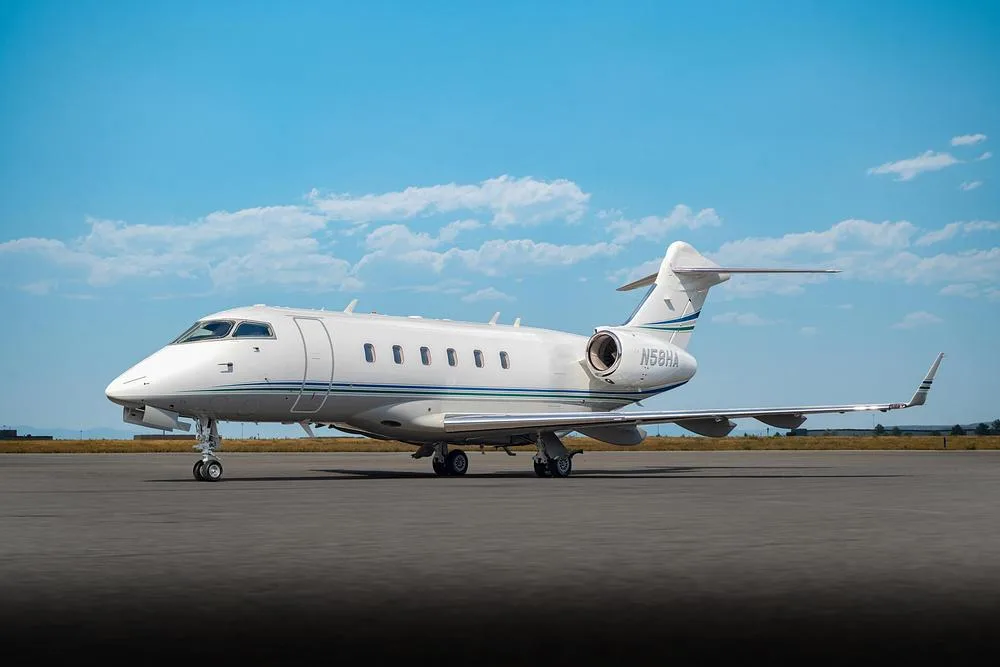 A white Bombardier Challenger 300 business jet with blue and green stripes, registration N58HA, parked on an asphalt tarmac under a blue sky.
