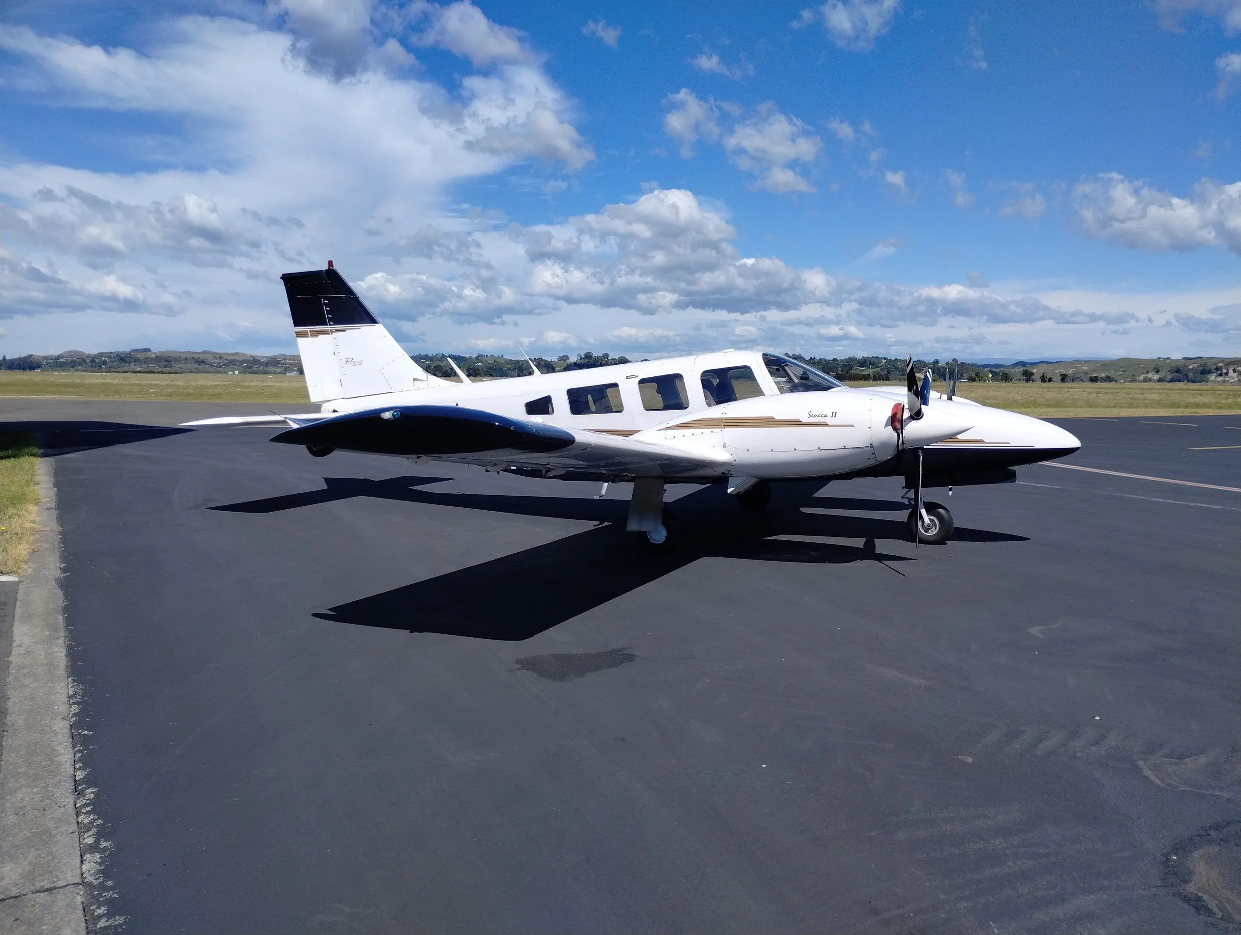 A white and black Piper Seneca II aircraft parked on an asphalt runway under a blue sky with clouds.