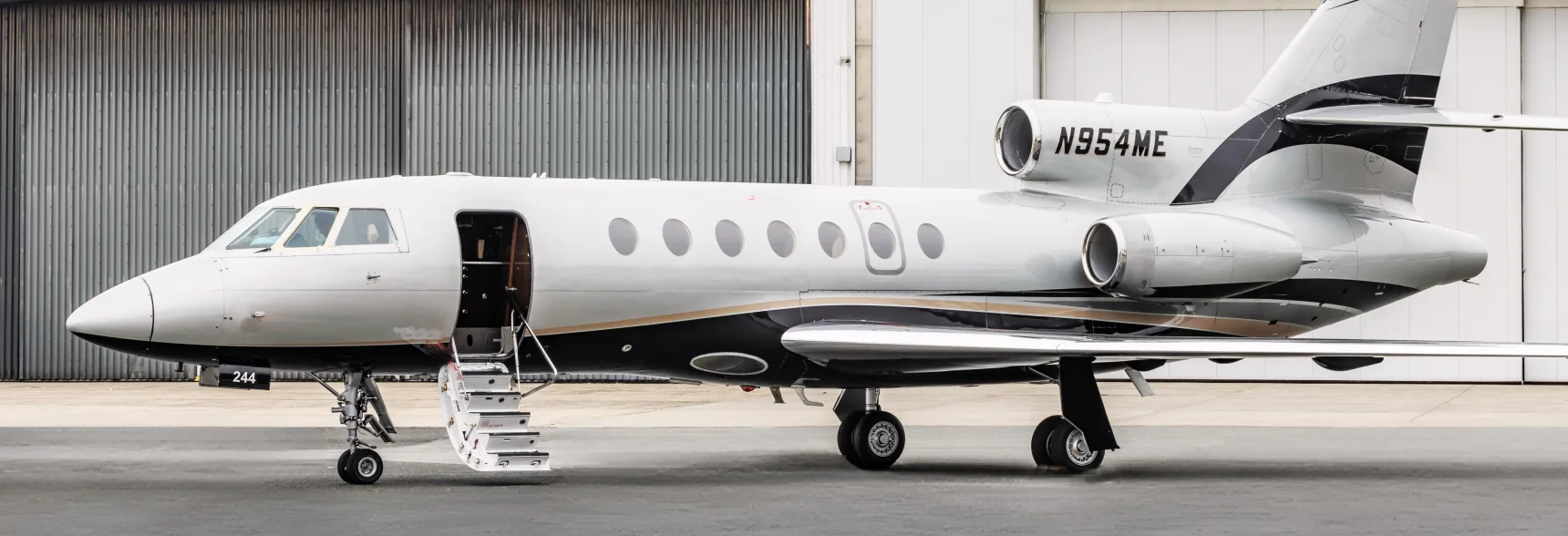 A close-up view of a silver and black Dassault Falcon business jet with gold accent stripes, parked on an apron with its airstairs deployed.