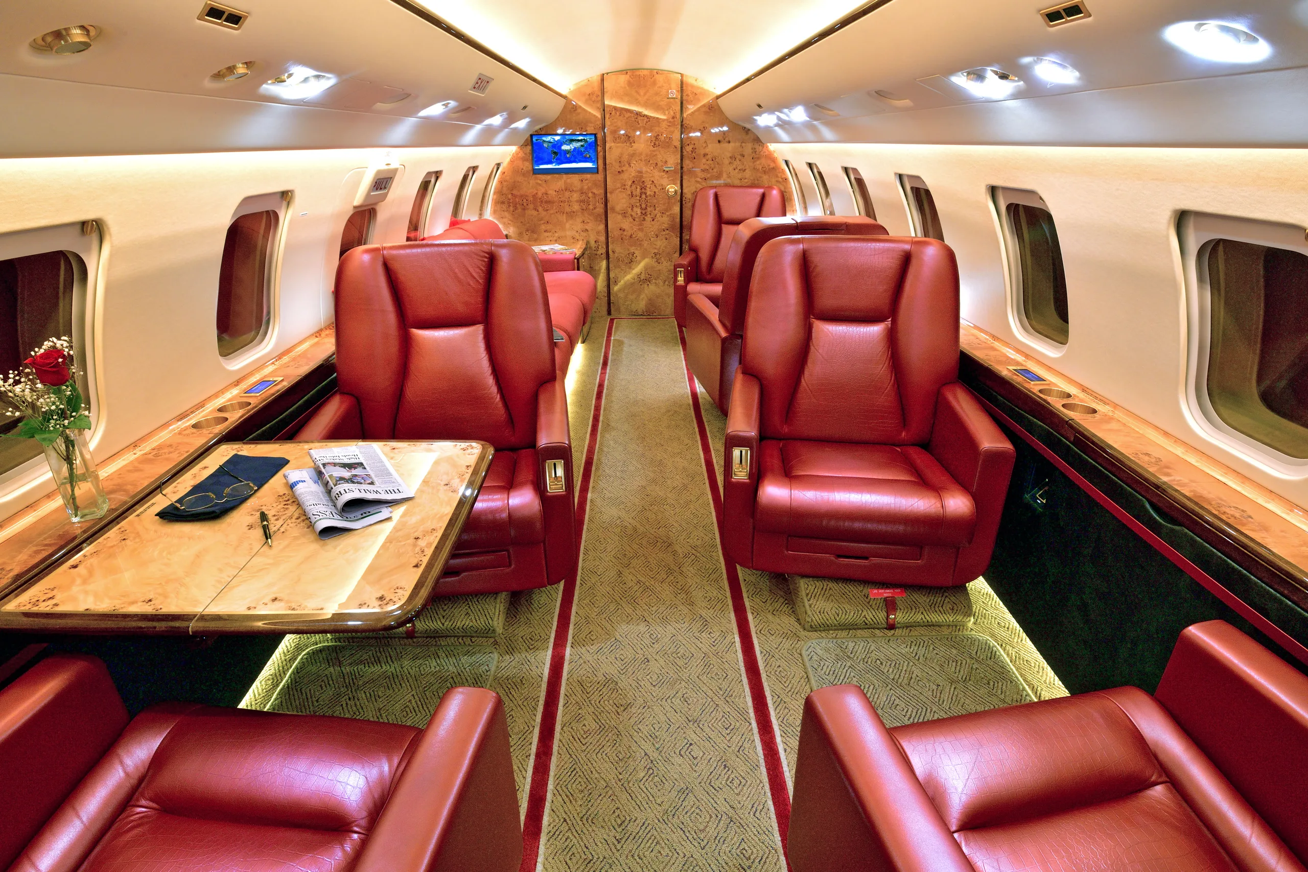 The luxurious cabin interior of a private jet with red leather seats, a wooden table, and a vase of red roses.