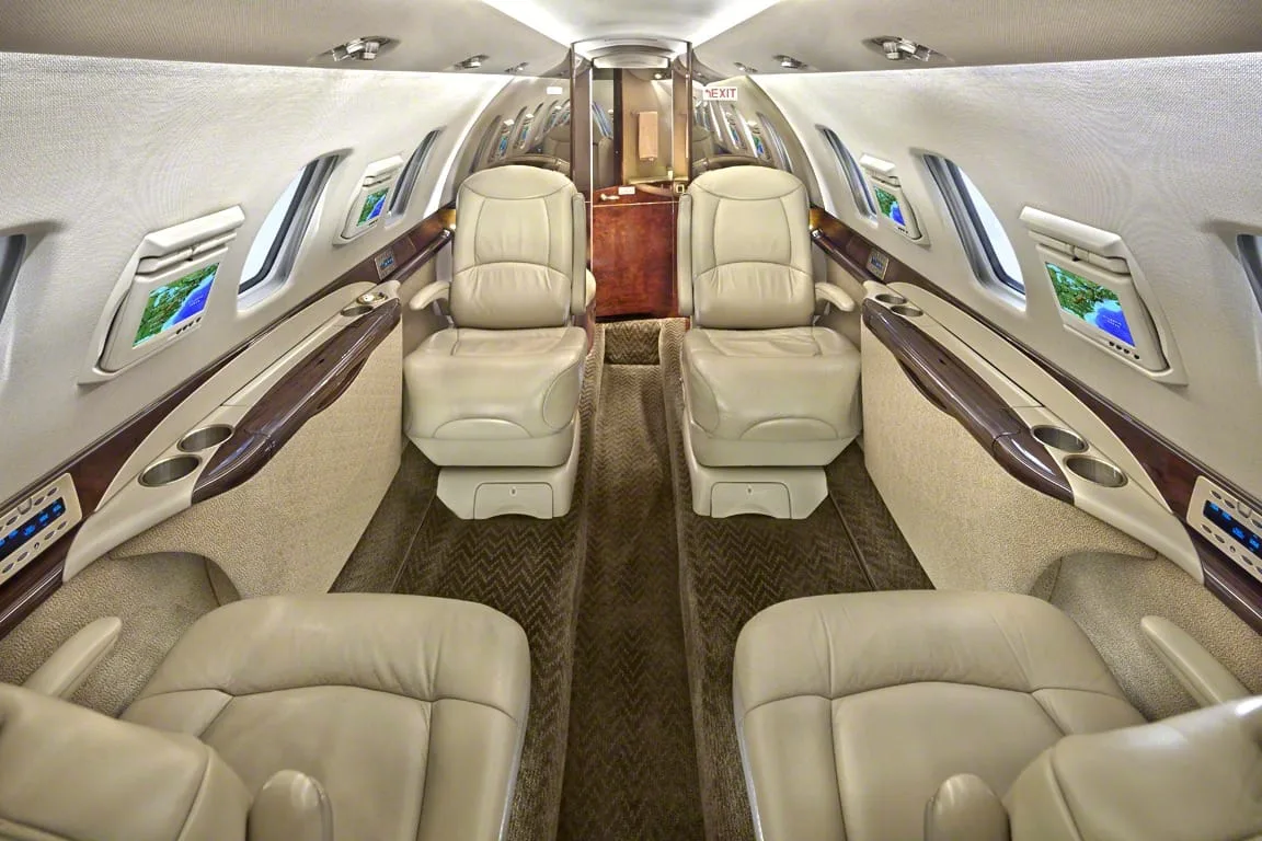 The luxurious interior cabin of a private jet with beige leather seats and dark wood trim.