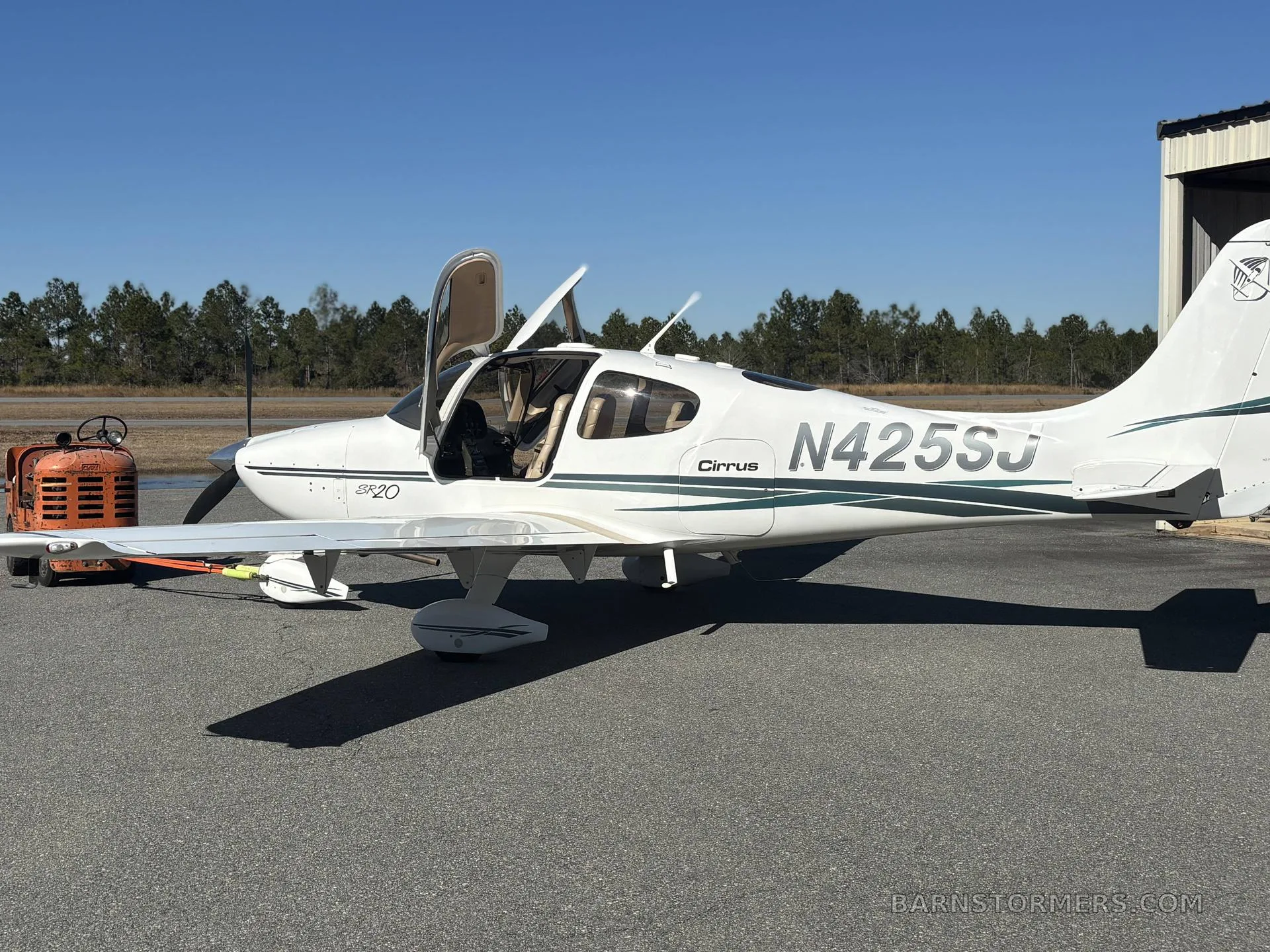 A white Cirrus SR20 aircraft with green stripes and tail number N425SJ is parked on an airfield with its doors open.