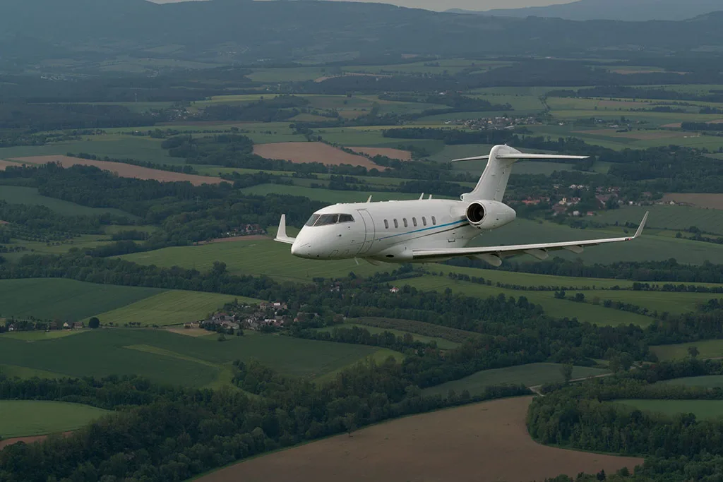 A white business jet with a blue stripe flying over a patchwork of green fields and forests under a cloudy sky.