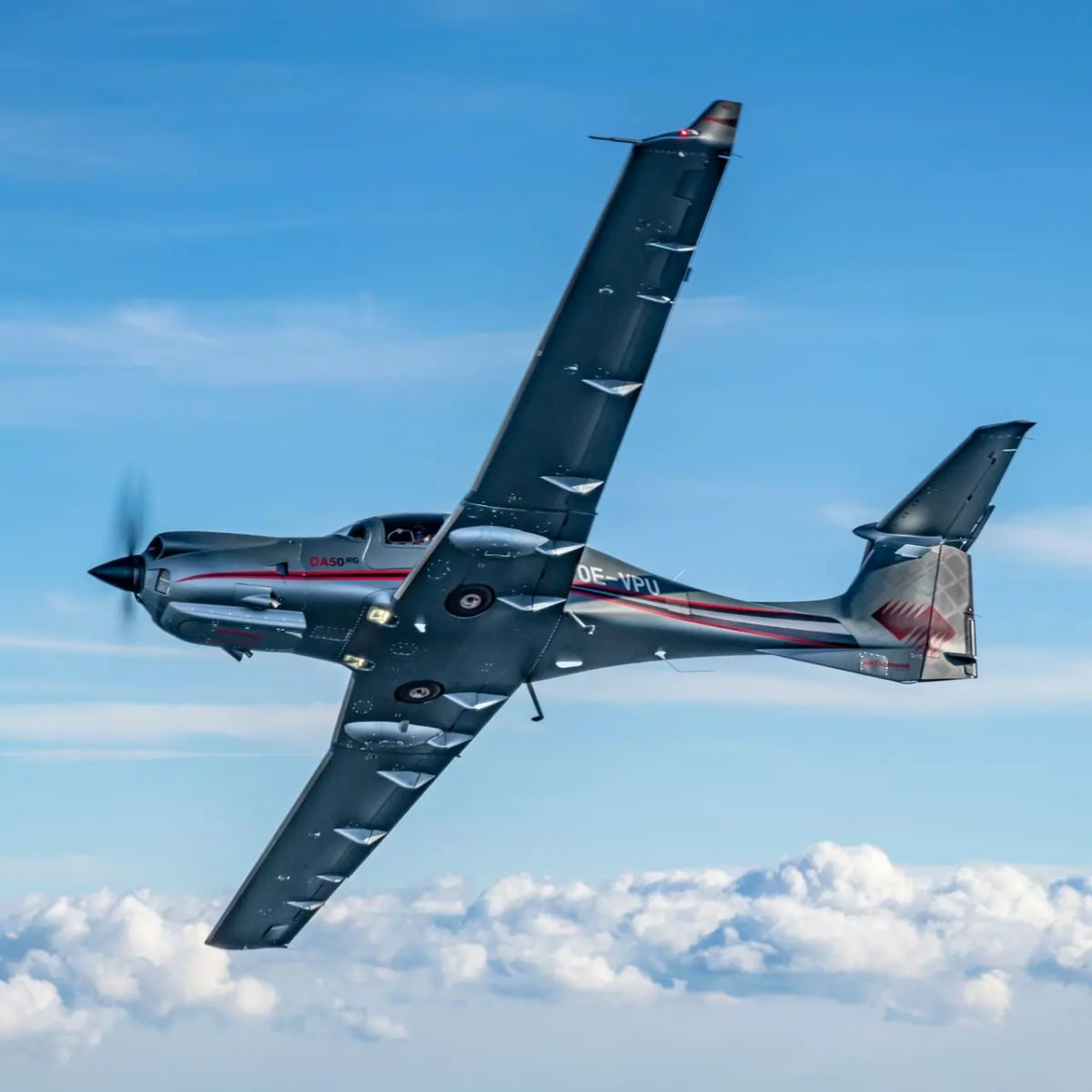 A grey Diamond DA50 RG single-engine aircraft with red stripes flying above a layer of white clouds.
