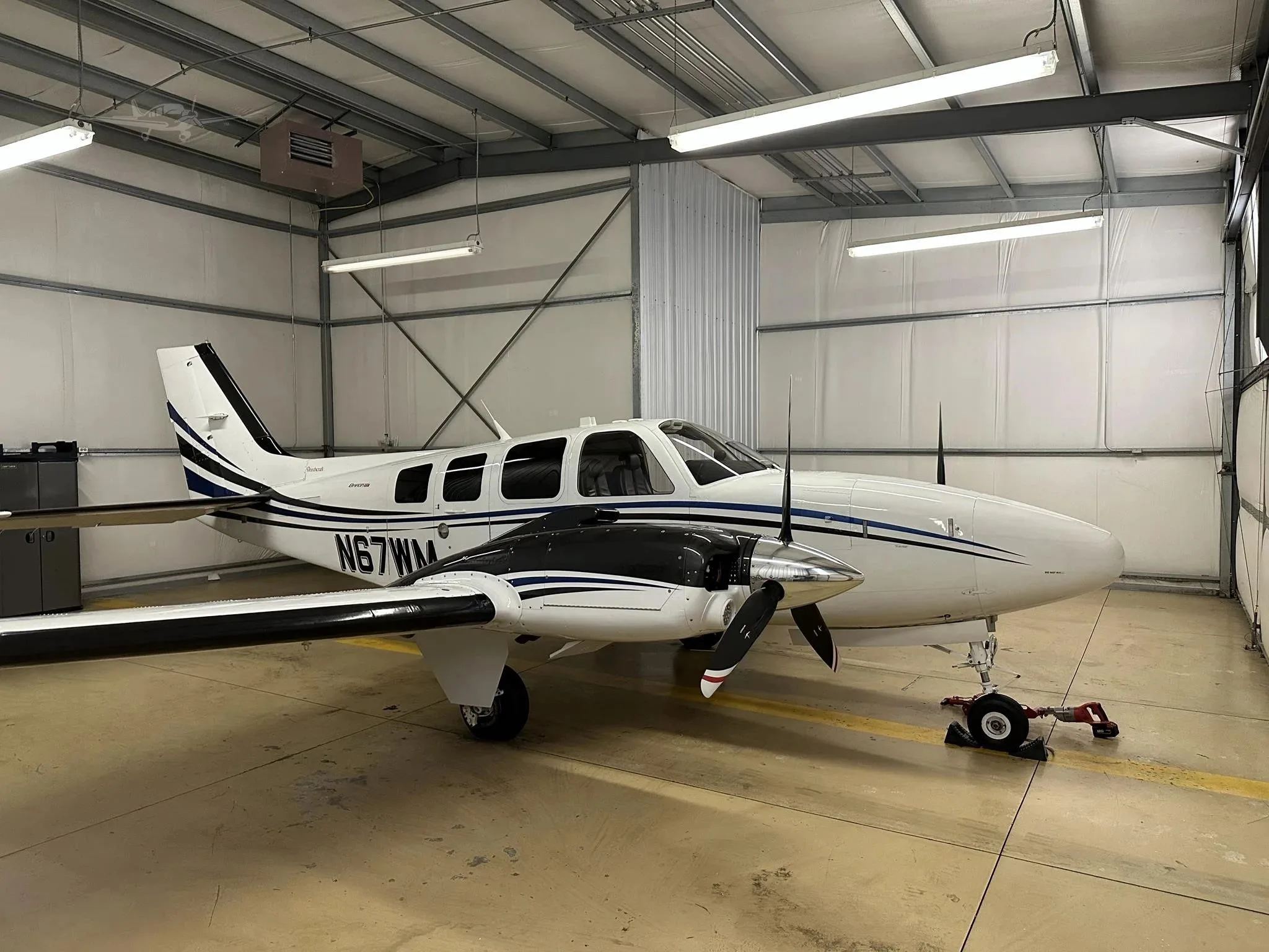 A white and blue Beechcraft Baron N67WM parked inside a hangar, viewed from the front-right.