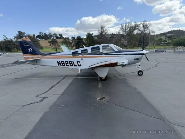 A white, blue, and copper Beechcraft Bonanza N926LC parked on a tarmac under a partly cloudy sky.