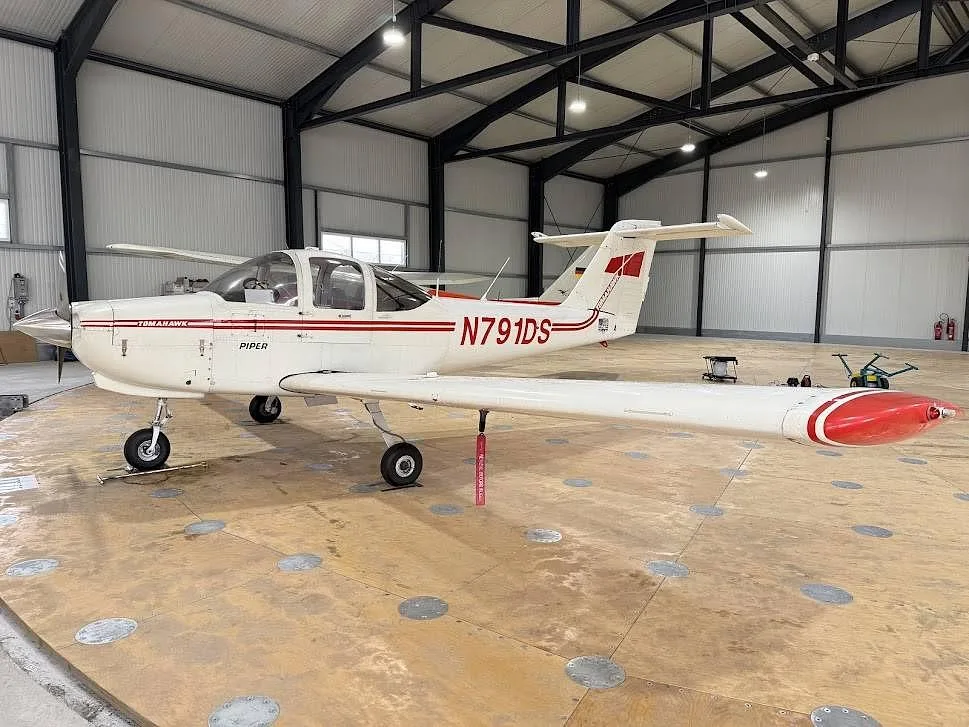 A white Piper Tomahawk aircraft with red stripes and registration N791DS is parked inside a hangar, viewed from the right side.