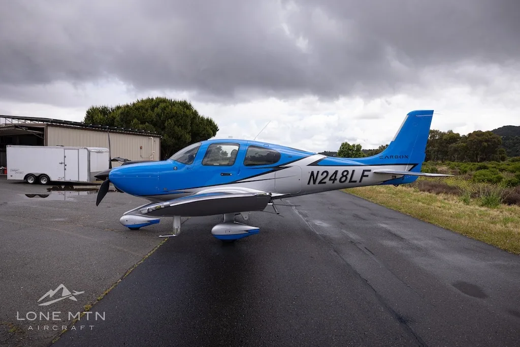 A blue and silver single-engine propeller aircraft (Cirrus SR22) with registration N248LF parked on a wet tarmac next to a hangar.