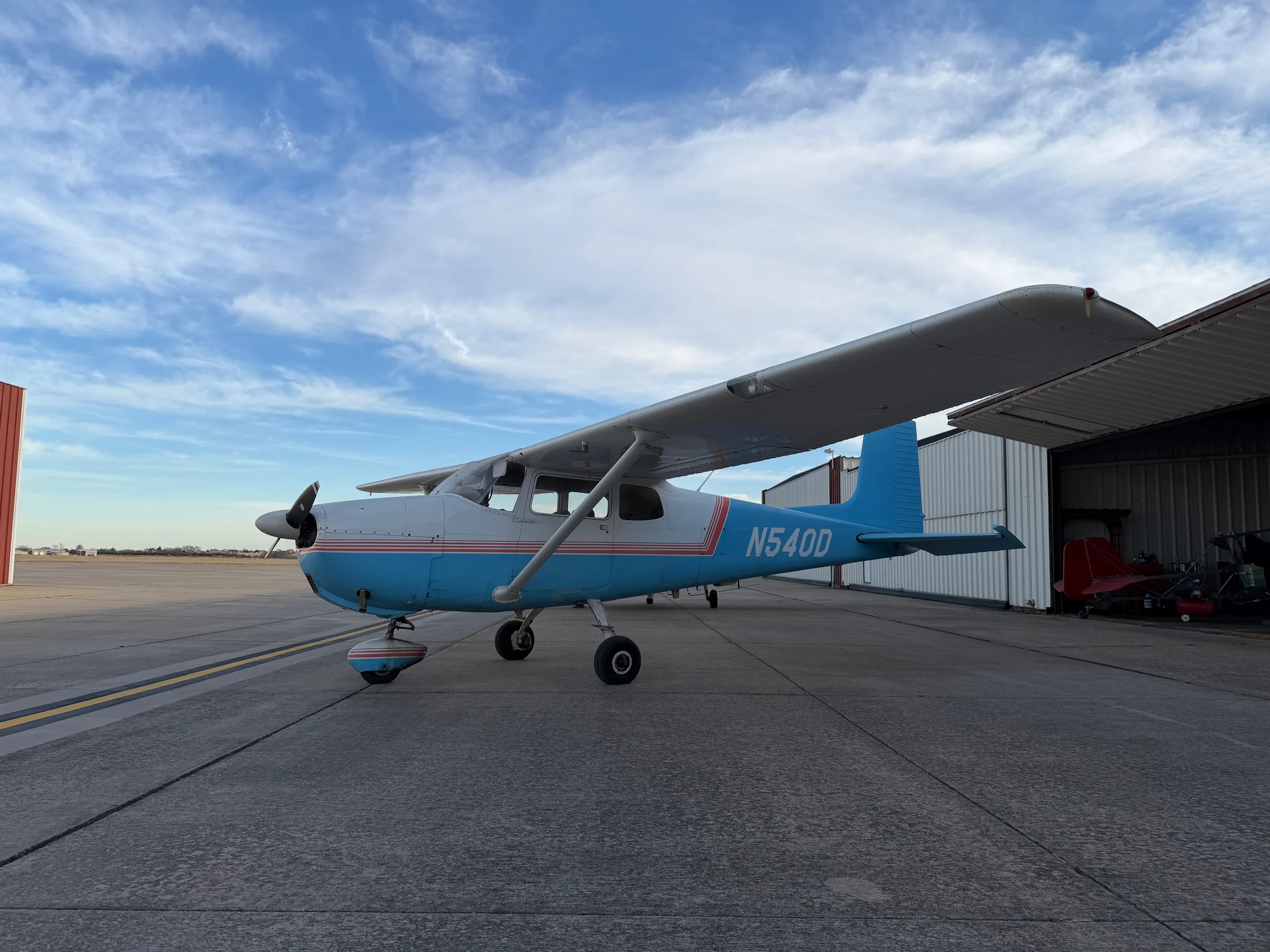 A white and light blue Cessna 172, registration N540D, parked on an airfield with hangars under a partly cloudy sky.