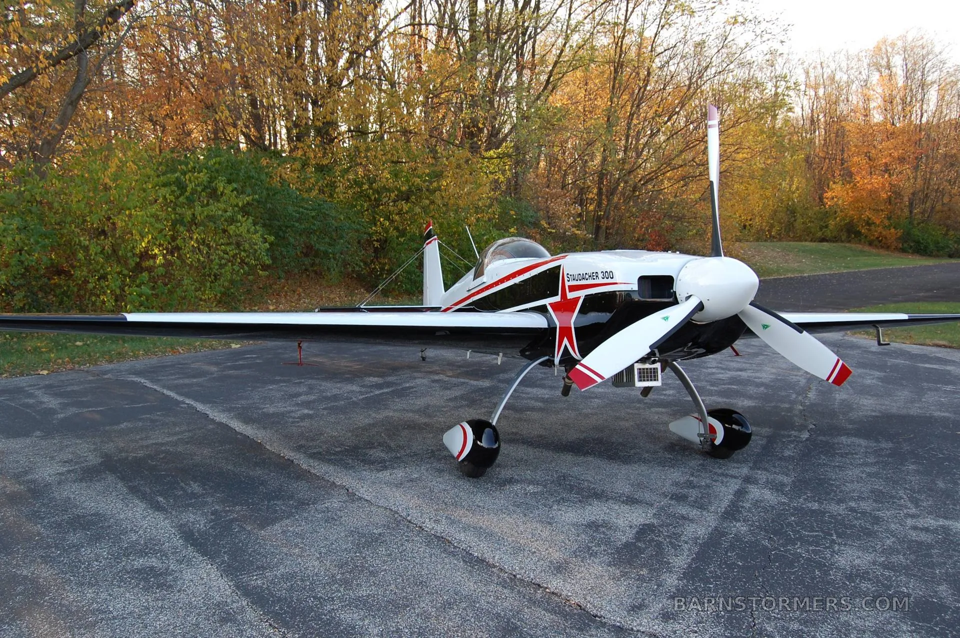 A white, black, and red Staudacher 300 aerobatic aircraft is parked on an asphalt surface with autumn trees in the background, viewed from the front-right.