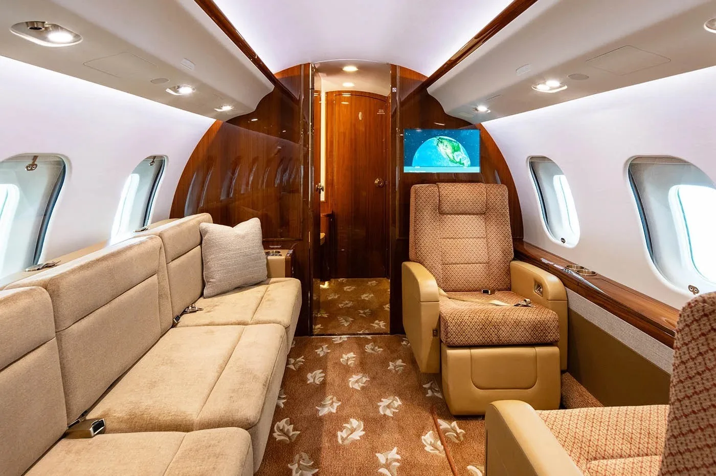 The aircraft cabin features a beige sofa, an individual seat, and a wall-mounted TV screen.