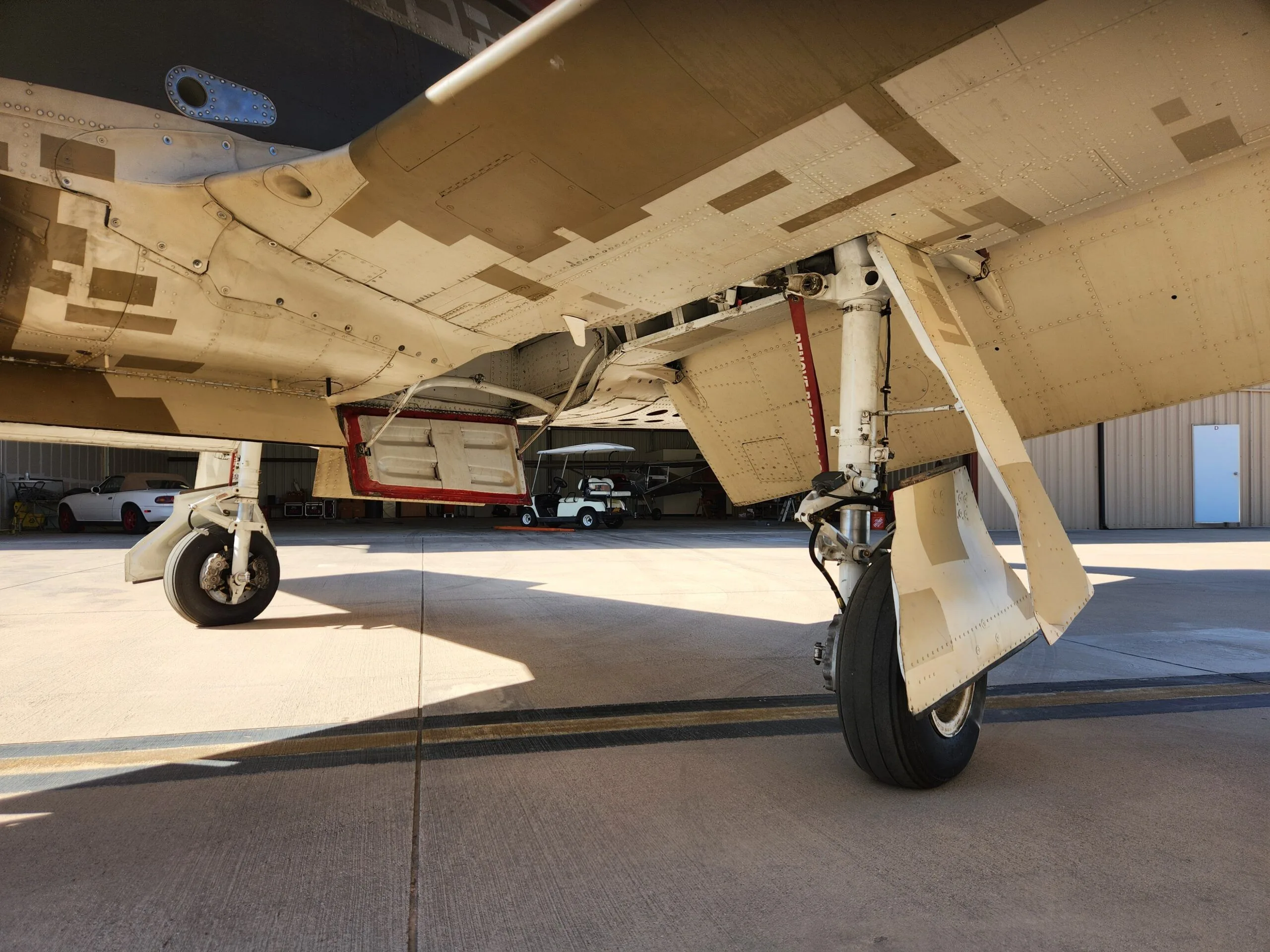 The underside of a T-28 Trojan aircraft's wing, showing the landing gear and a storage compartment, parked on an airfield.