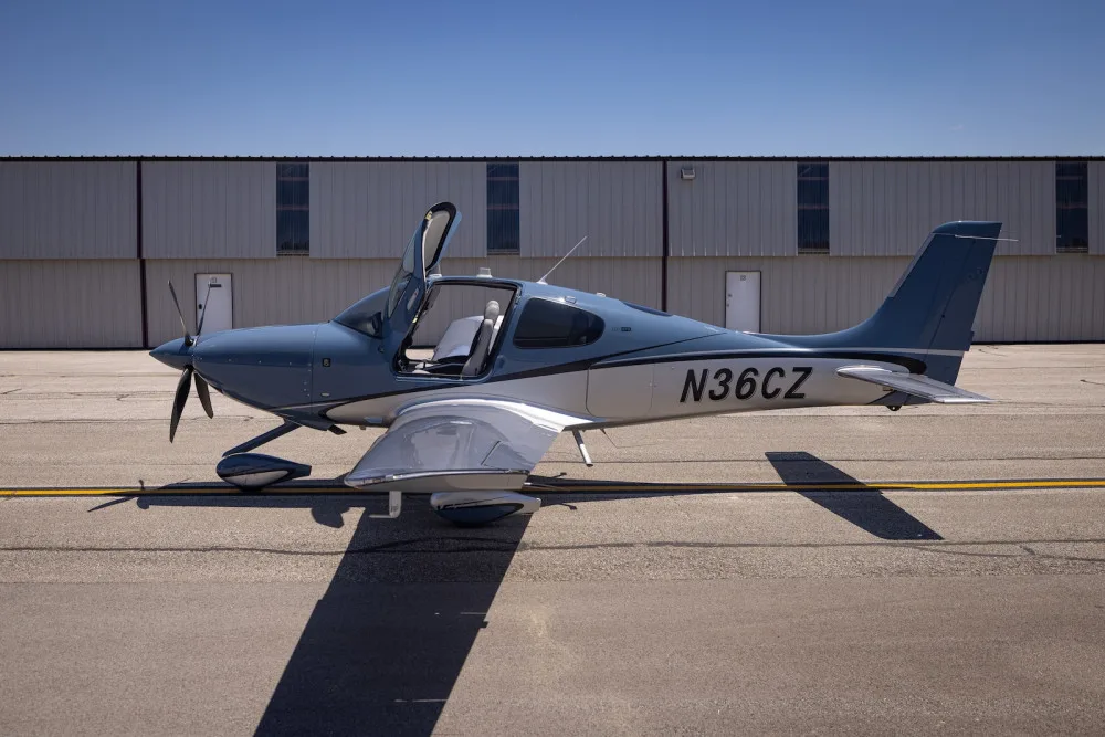 A blue and silver Cirrus SR22T aircraft with open doors parked on an airport tarmac, viewed from the side.