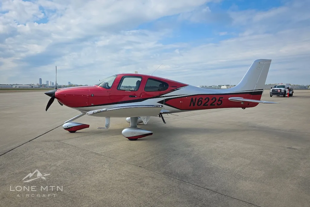 A red and silver Cirrus SR22 aircraft, N6225, parked on an airport apron with a city skyline in the distance.