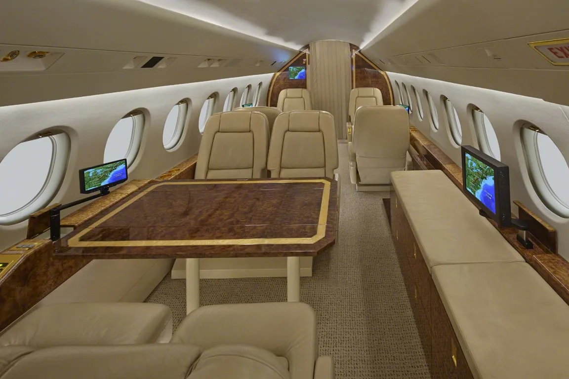 A wide view of a private jet's cabin with beige leather seating, two polished wood tables, and individual entertainment screens.