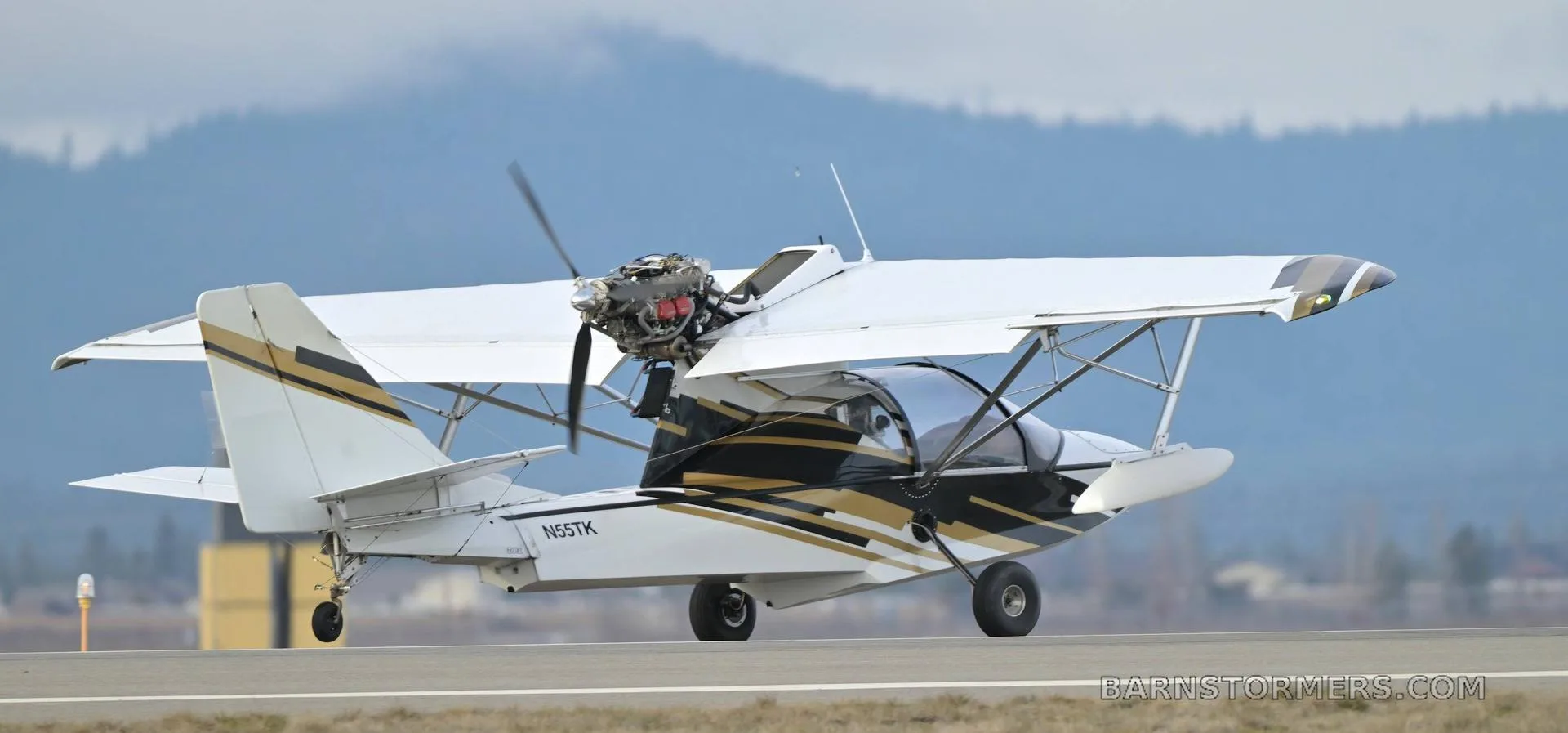 A white and gold amphibious light sport aircraft with floats and wheels on a runway with mountains in the background.