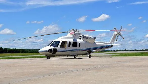 A white Sikorsky S-76 helicopter with blue stripes and red rotor tips, parked on an asphalt tarmac under a blue sky with clouds.