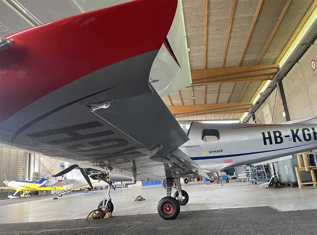 Underside view of the red-tipped wing and main landing gear of a silver Sonaca 200 aircraft in a hangar.