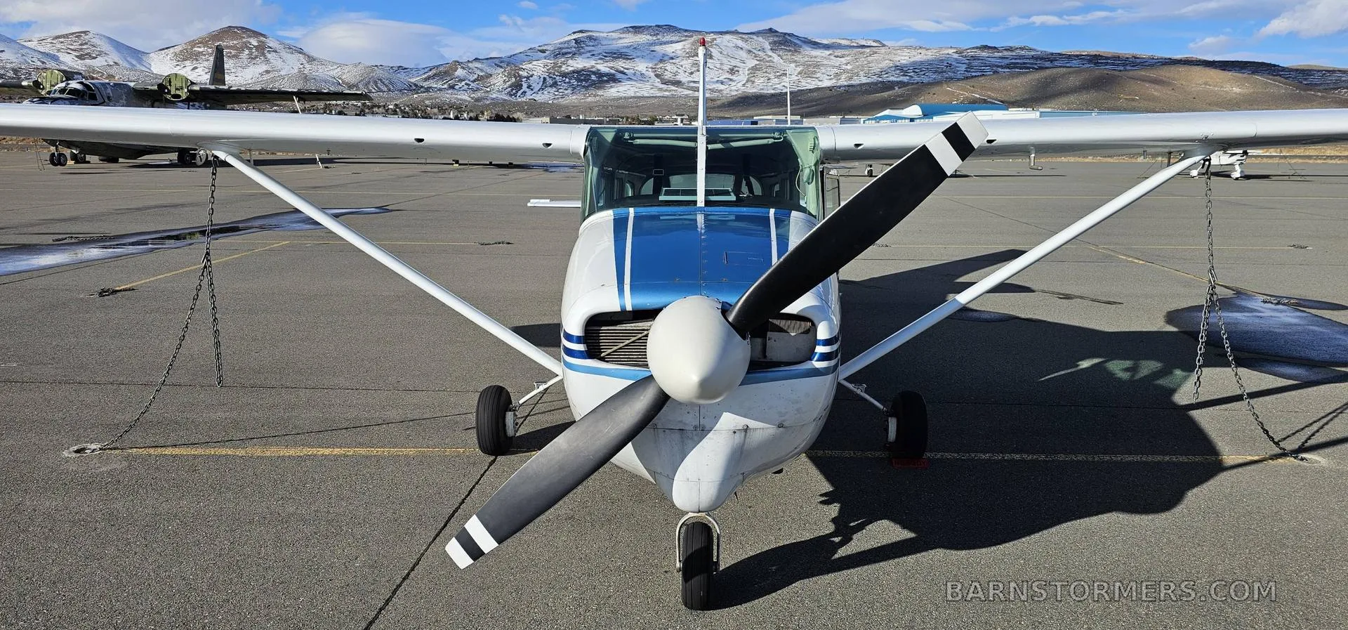 A head-on view of a white and blue Cessna 182 aircraft with its propeller, parked on an airfield with snowy mountains.