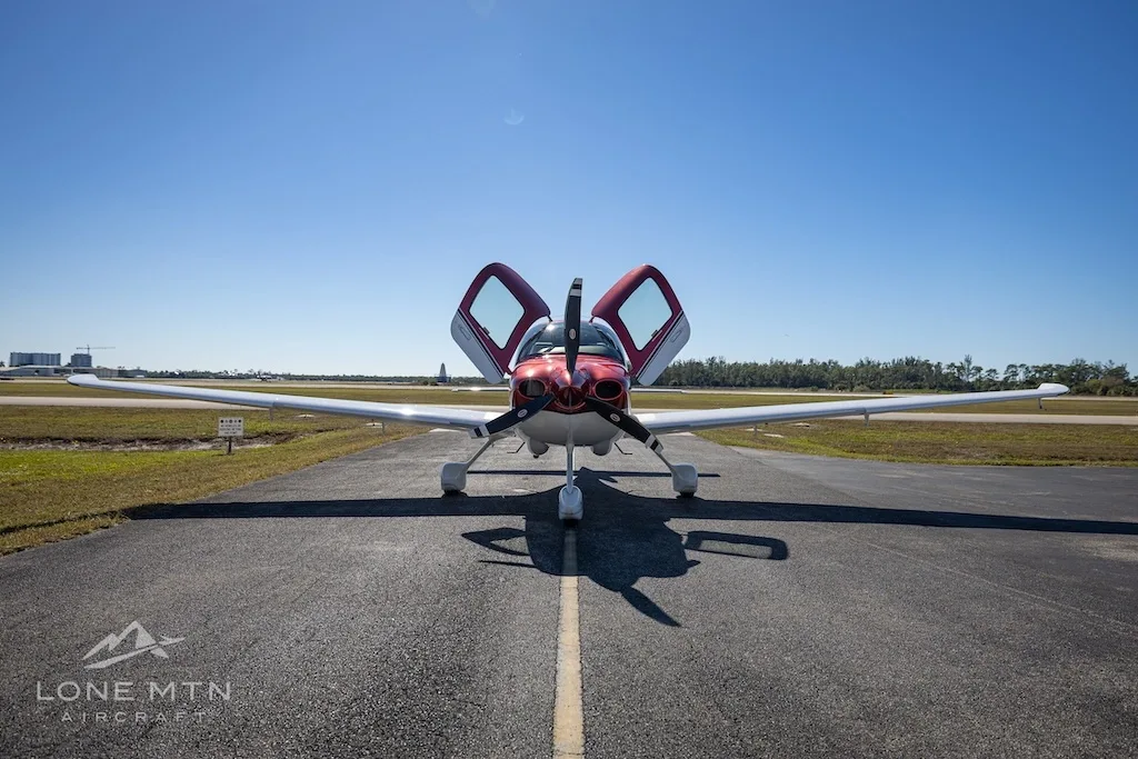 A head-on view of the red and white Cirrus SR20 aircraft (N432WB) with its doors open, parked on an airport runway.