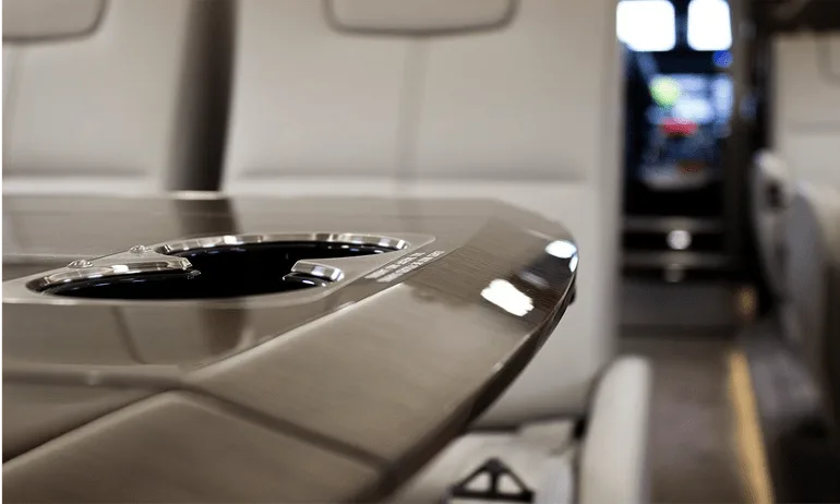 A close-up of a polished table with cup holders inside an aircraft cabin, with seats and the cockpit visible in the background.